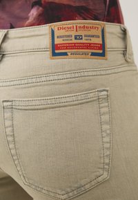 Jeans de mezclilla beige claro con un bolsillo trasero y una etiqueta rectangular marrón con detalles en azul y rojo, que dice "Diesel Industry."