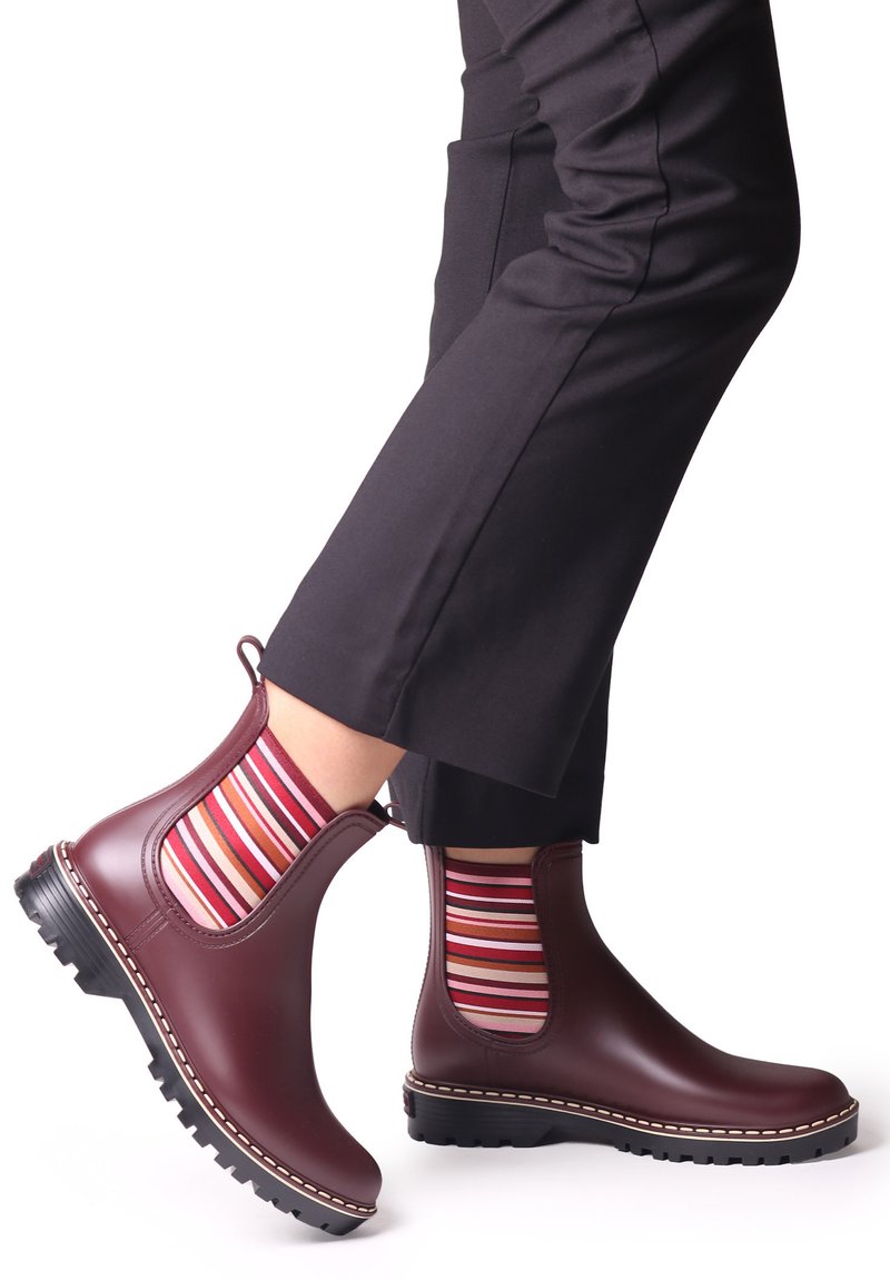 Bottines Chelsea en caoutchouc bordeaux avec des panneaux latéraux élastiques, semelle crantée noire et couture contrastante, associées à des chaussettes rayées multicolores.