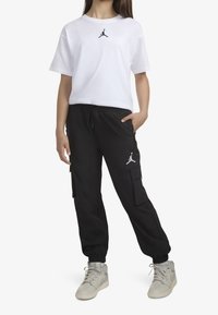 Biała koszulka z czarnym logo Jordana; czarne spodnie cargo z bocznymi kieszeniami i sznurkiem; szare buty sportowe z sznurówkami.