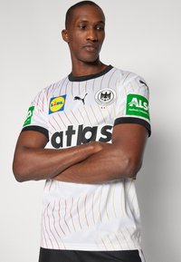 Bijela sportska majica s crvenim i žutim dijagonalnim prugama. Sadrži logotipe Lidl i Alsid te crni ovratnik s natpisom "atlas" u podebljanom fontu.