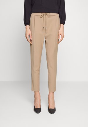 Vrouw draagt beige taps toelopende broek met trekkoord in de taille en zwarte puntige hakken, staand tegen een lichtgrijze achtergrond.