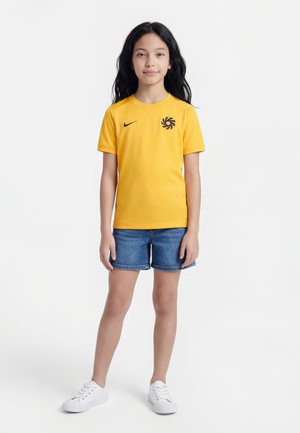 Fată cu păr lung și închis la culoare, purtând un tricou galben Nike cu un logo negru, pantaloni scurți din denim albastru și adidași albi, stând pe un fundal alb.