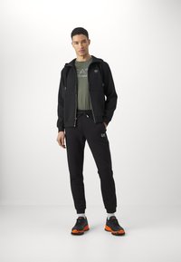 Felpa nera con zip e due tasche frontali, t-shirt verde con logo, pantaloni sportivi neri e arancioni, e sneakers nere e arancioni. Tessuto morbido, vestibilità casual.