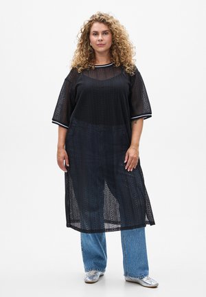 Tunique en dentelle noire avec une coupe ample, des manches courtes et une bordure rayée, portée par-dessus un jean large bleu clair et des chaussures argentées.