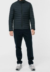 Černá péřová bunda s horizontálním prošíváním, zapínáním na zip, vysokým límcem, v kombinaci s tmavými manšestrovými kalhotami a šedými teniskami.