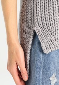 Ljusgrå stickad tröja med ribbad textur, med en sidoklyfta, bärs över slitna blå denimjeans.