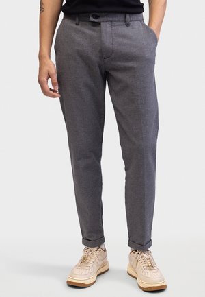 Homme portant un pantalon gris texturé slim avec les revers roulés et des baskets beige, debout les mains dans les poches.