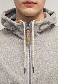Felpa grigia con zip e colletto alto, cordini grigi e dettagli in pelle beige. Presenta una cerniera metallica e un tessuto testurizzato.