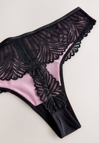 Čierne čipkované tangá s prepracovanými kvetinovými a geometrickými vzormi na svetlom mauve textilnom pozadí.