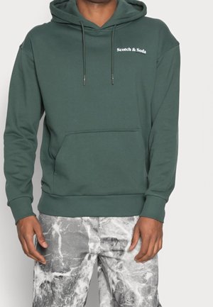Mann trägt einen dunkelgrünen Hoodie mit Fronttasche und weißem "Scotch & Soda"-Logo, kombiniert mit grau-weißen marmorierten Hosen.