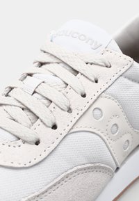 Bílá teniska s texturovaným semišem a síťovinovými materiály. Má výrazné logo s kruhovými výřezy a ploché tkaničky. Minimalistický design.