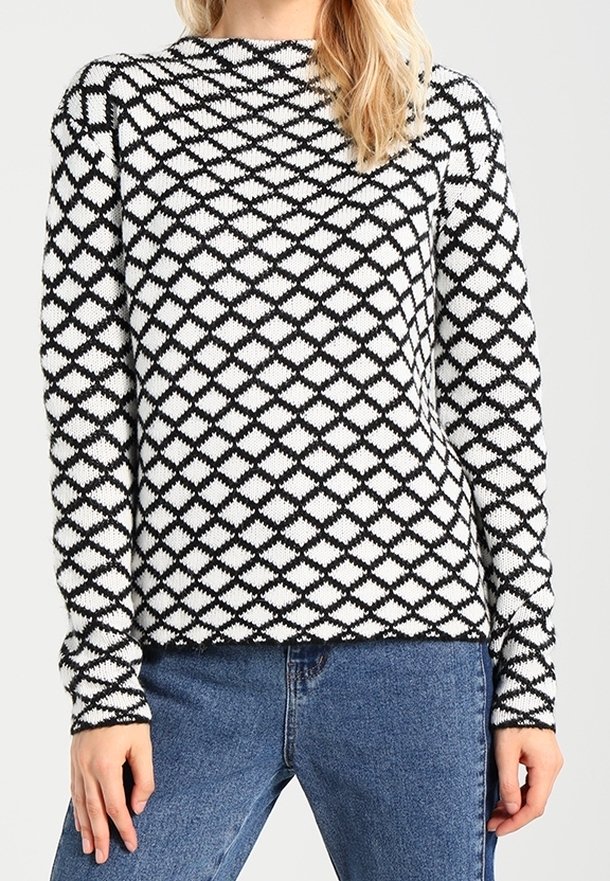 Femme portant un pull à manches longues à motifs en losange noir et blanc, associé à un jean bleu, sur un fond uni.
