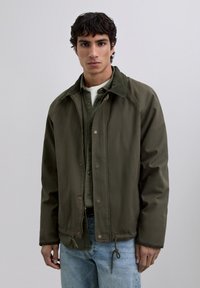 Young man with short curly hair wearing an olive green jacket over a green button-up shirt and light blue jeans against a plain background.