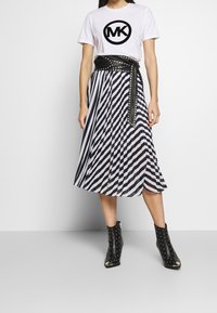 Kvinna som bär en vit t-shirt med svart "MK"-logga, svart nitarbälte, svart och vit randig plisserad kjol och svarta nitarstövletter.