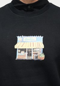 Μικρή πολύχρωμη εικονογράφηση μιας βιτρίνας με το όνομα Καρλίτος, με τέντα, ανοιχτή πόρτα, ράφια και μια γλάστρα, σε μαύρο μπλουζάκι.