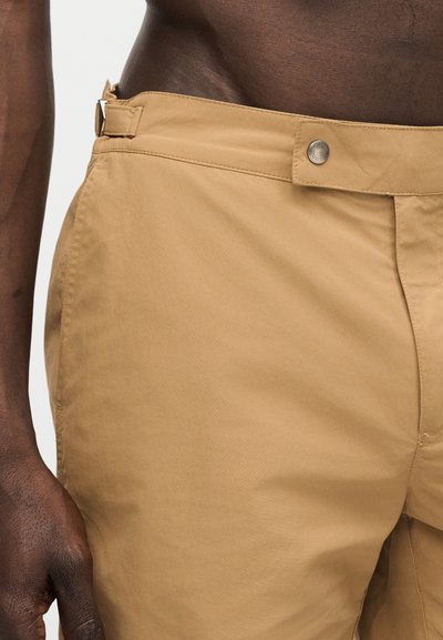 Közeli kép egy sötét bőrű személyről, aki bézs nadrágot visel, gombbal és oldalsó állító csattal, kezét lazán oldalt tartva.