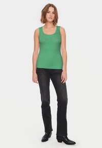 Canottiera verde realizzata in tessuto elasticizzato con scollo a giro, abbinata a jeans scuri e stivali neri alla caviglia.