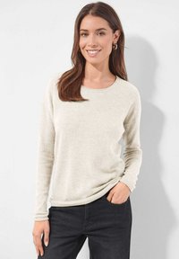 Junge Frau mit langen braunen Haaren, die ein hellbeiges Langarmoberteil und schwarze Hosen trägt. Sie steht mit einer Hand in der Tasche und lächelt.