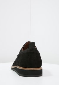 Scarpe eleganti nere in suede viste da dietro, con una suola intermedia marrone, suola nera e una linguetta sul tallone, posizionate su una superficie bianca.