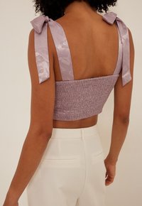 Cropped top lilla con texture smockata e ampie spalline in satin. La schiena è aperta, abbinato a pantaloni bianchi a vita alta.