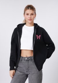 Sweat à capuche zippé noir avec motif papillon rose sur la poitrine gauche, associé à un t-shirt court blanc et un jean gris taille haute.