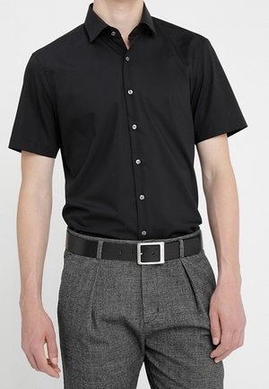 Homme portant une chemise à manches courtes noire ajustée, rentrée dans un pantalon texturé gris avec une ceinture noire et une boucle en argent.