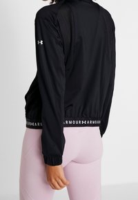 Kvinna som bär en svart långärmad träningsjacka med meshnätpaneler och ljusrosa tajta leggings, sedd bakifrån.