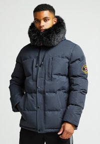 Jūros mėlynumo pūkinė striukė su dideliu dirbtinio kailio apykakle, dviem priekinėmis kišenėmis, matoma siuvimo linija ir logotipo pleistru ant rankovės.