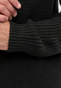 Zwarte gebreide trui met ribbelstructuur, close-up van de mouw, die de flexibele pasvorm en naadloze ontwerpelementen laat zien.