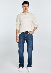 Beige henley-shirt med lange ærmer og tre brune knapper, kombineret med mørkeblå jeans med lige ben og hvide sneakers. Simpelt design, glat tekstur.