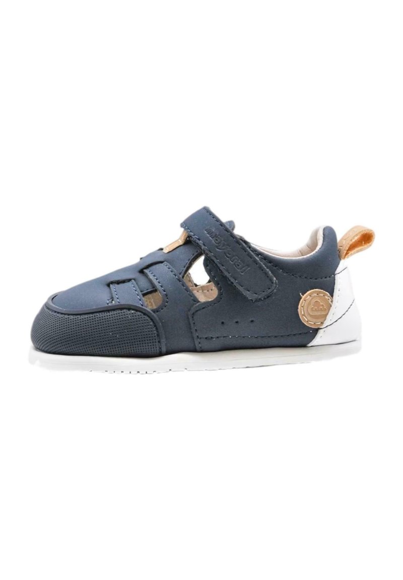 Zapato para bebé de color azul marino con un diseño transpirable, correa de velcro, suela blanca y acento de cuero marrón. Material texturizado con características recortadas.