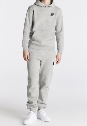 Ensemble en sweat-shirt et pantalon de jogging gris en tissu doux, doté d'une capuche et d'un logo sur la poitrine, associé à des baskets blanches.