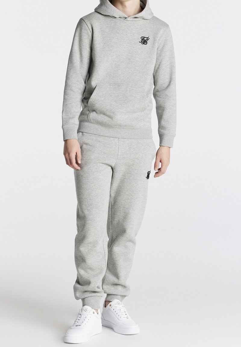 Grau Sweatshirt und Sweatpants-Set aus weichem Material, mit Kapuze und Logo auf der Brust, kombiniert mit weißen Sneakers.
