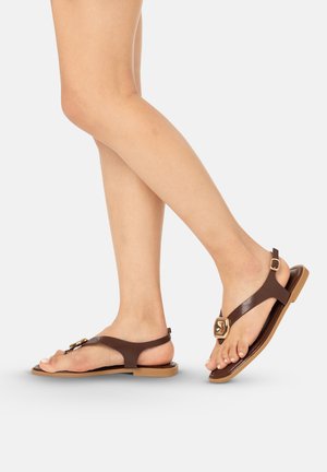 Pies con sandalias planas de cuero marrón con detalles de hebilla dorada sobre un fondo claro, un pie ligeramente levantado.