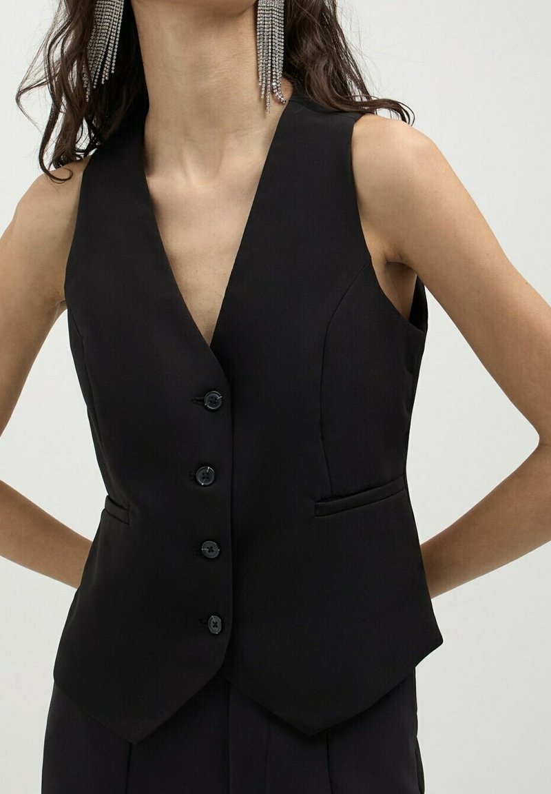 Svart ärmlös väst med figursydd design, fem knappar och två framfickor. Mjuk textur och skarp, triangulär halsringning.