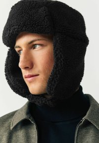 Black fleece trapper hat with ear flaps, textured fabric, rounded top, and adjustable chin strap. Worn with a dark turtleneck and patterned jacket.