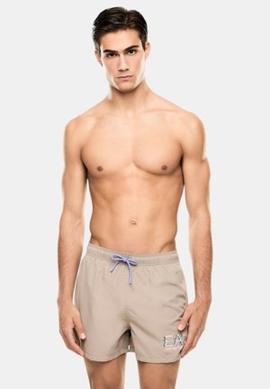 Giovane uomo con capelli scuri e corti in piedi, senza maglietta, indossa pantaloncini da bagno beige con coulisse blu e logo sulla gamba sinistra.