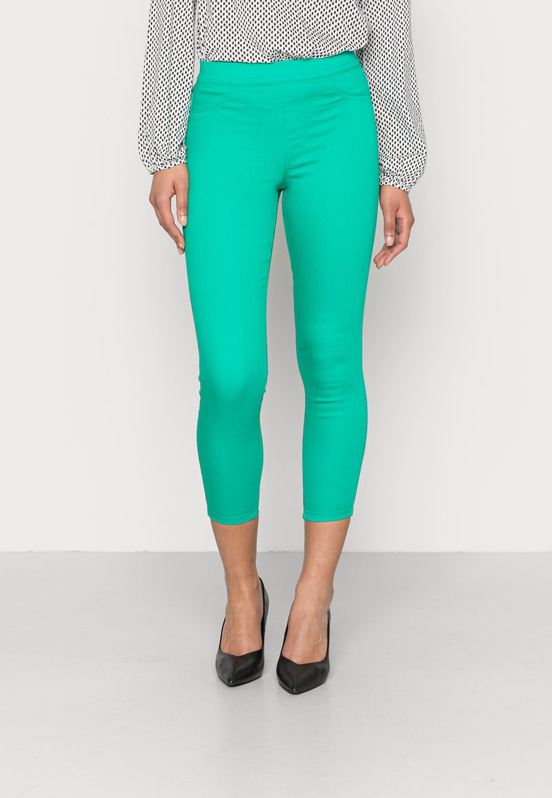 Calças capri em turquesa, feitas de um tecido elástico com um design ajustado, combinadas com sapatos de salto agudo pretos e uma blusa de bolinhas.