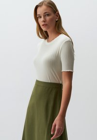 Biała, prążkowana bluzka z krótkim rękawem i falbaniastym dekoltem, zestawiona z długą oliwkową spódnicą o gładkiej fakturze i dopasowanym kroju.