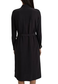 Czarna sukienka z długim rękawem, z kołnierzykiem, paskiem w talii i subtelnym połyskiem; ma prosty krój i zapięcia na guziki przy rękawach.