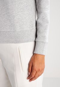 Pullover grigio in cotone con polsini a costine, caratterizzato da una superficie strutturata e un design aderente, abbinato a pantaloni bianchi con dettagli a cerniera laterale.