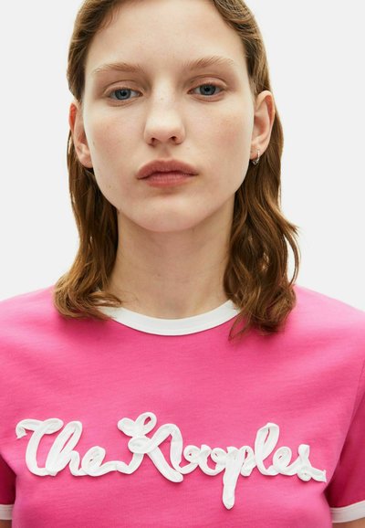 Roze katoenen t-shirt met een witte geribbelde kraag, voorzien van geprofileerde witte letters die "The Krooples" op de borst spellen.