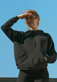 Schwarzer Kapuzenpullover aus weichem Stoff mit einer Fronttasche und eingeprägtem Logo. Das Model steht vor einem blauen Himmel.