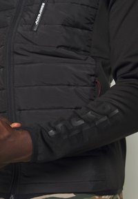 Black padded jacket with a smooth, stretchy sleeve featuring embossed logo. Zippered front pocket and design contrast on materials.