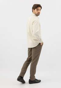 Camicia beige chiaro a maniche lunghe con dettagli in bottoni, abbinata a pantaloni marroni slim-fit e scarpe nere, mostrata di lato.