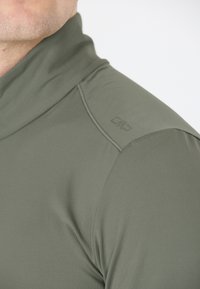 Olivengrøn sportsbluse med høj krave, glat tekstur og diskret logo på skulderen. Har buede sømme for en tætsiddende design.