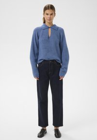 Modra pletena jopica s ovratnikom in dekoltejem v obliki ključavnice, združena z temno modrimi ravnimi hlačami in črnimi vezalkami. Gladka tekstura.