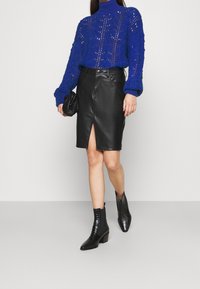 Modrý pletený svetr s vysokým límcem, černá kožená sukně tužkového střihu s předním rozparkem a černé kotníkové boty s texturovaným povrchem.