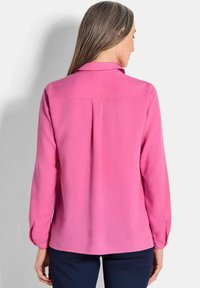 Camicia a maniche lunghe rosa con colletto, texture liscia e dettaglio a pieghe sul retro. I polsini hanno chiusure a bottoni. Indossata con pantaloni scuri.