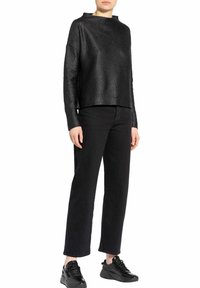 Mujer vestida con una camiseta negra de manga larga y textura, pantalones negros rectos y zapatillas negras, de pie sobre un fondo blanco.